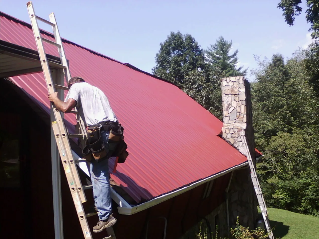 Roofer on a ladder beside a red metal roof and stone chimney — Soffit & Fascia Repair by Roofing Brothers in Gratis